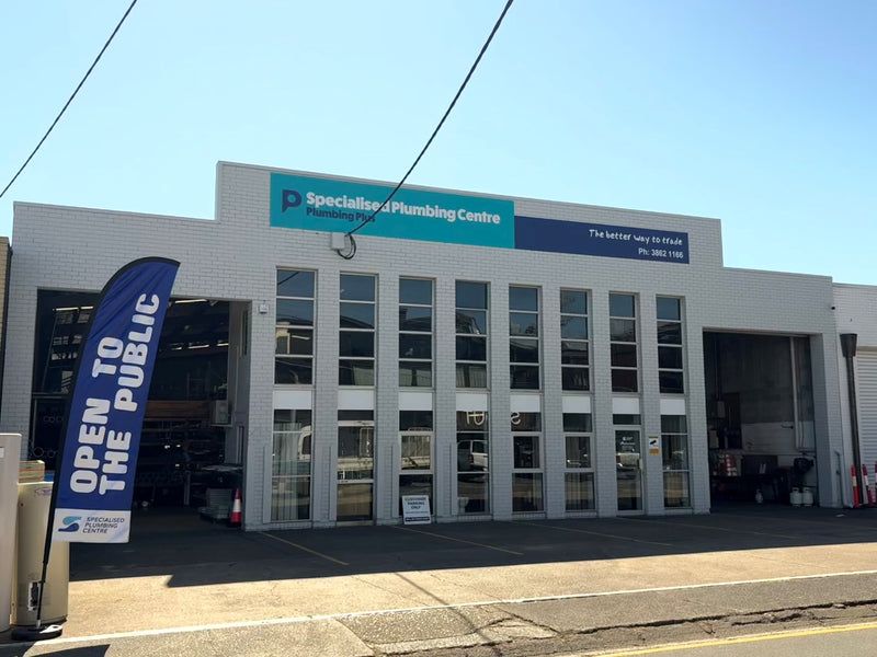 The exterior of the Specialised Plumbing Centre Albion Store under a clear blue sky, featuring a white brick facade, large garage doors, and a blue vertical banner that reads 'OPEN TO THE PUBLIC'.