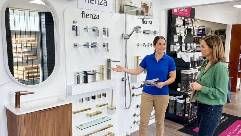 A showroom interior featuring Fienza bathroom fixtures, with a woman in a blue polo shirt gesturing toward wall-mounted fixtures while another woman stands nearby. Visible products include a white sink with a wood-look cabinet, a wall-mounted shower, and various faucets and accessories.